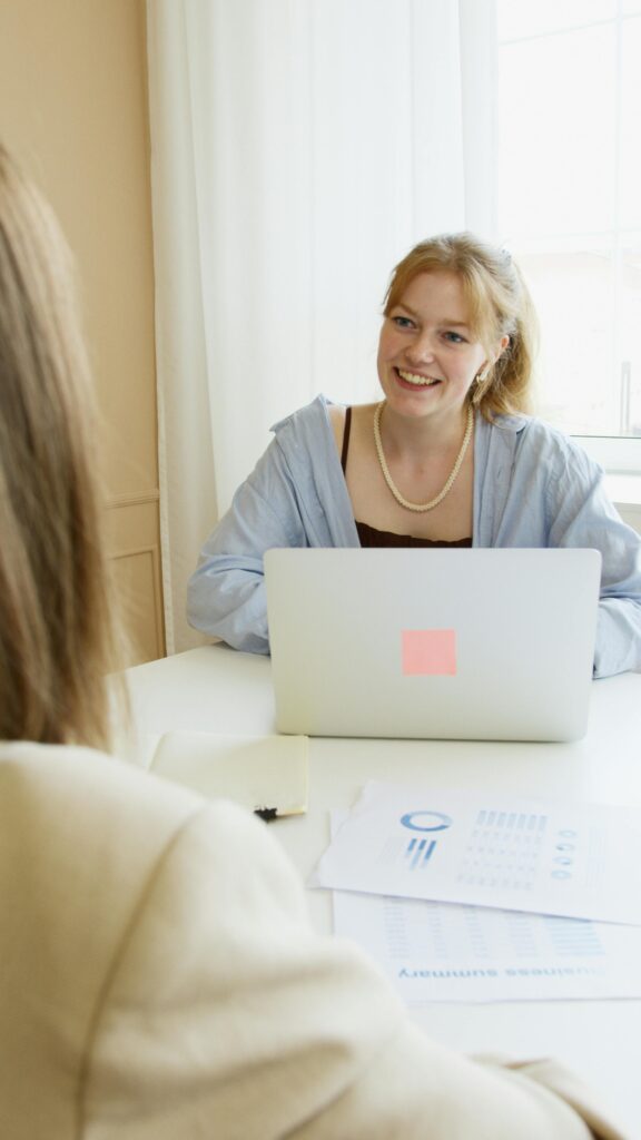 A young woman with a laptop smiling at another person in an indoor business setting.