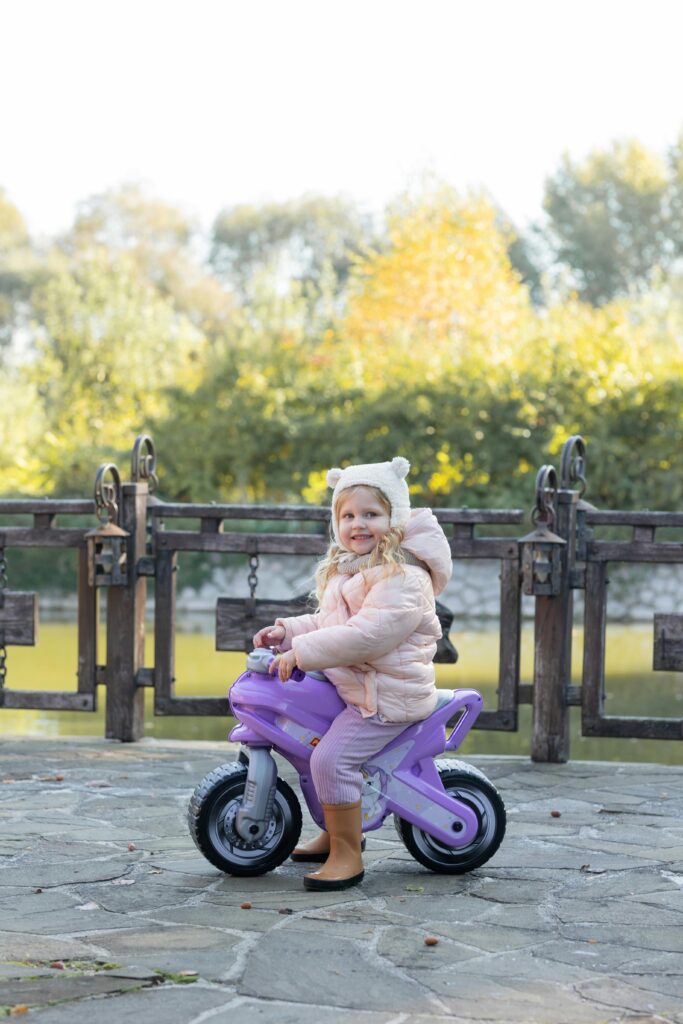 Happy young girl riding a purple toy motorcycle in a beautiful autumn park setting.